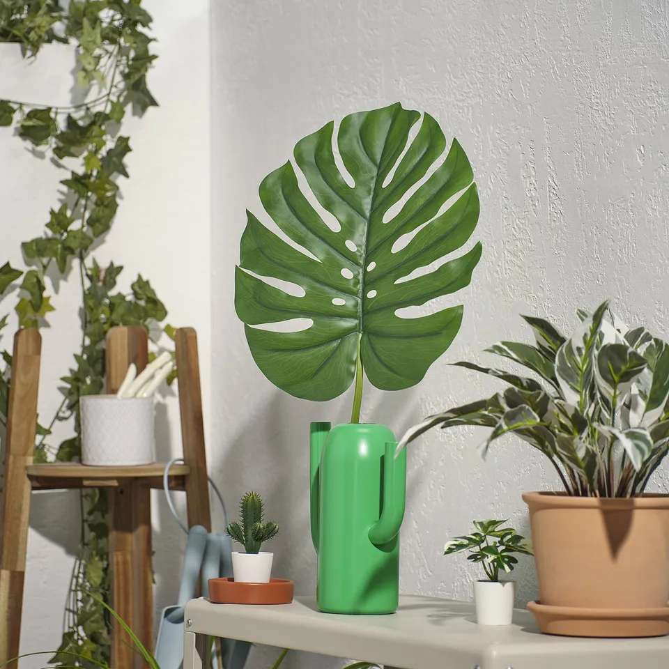 Large green leaf wall decor, made from recycled plastic, surrounded by various potted plants on shelves.