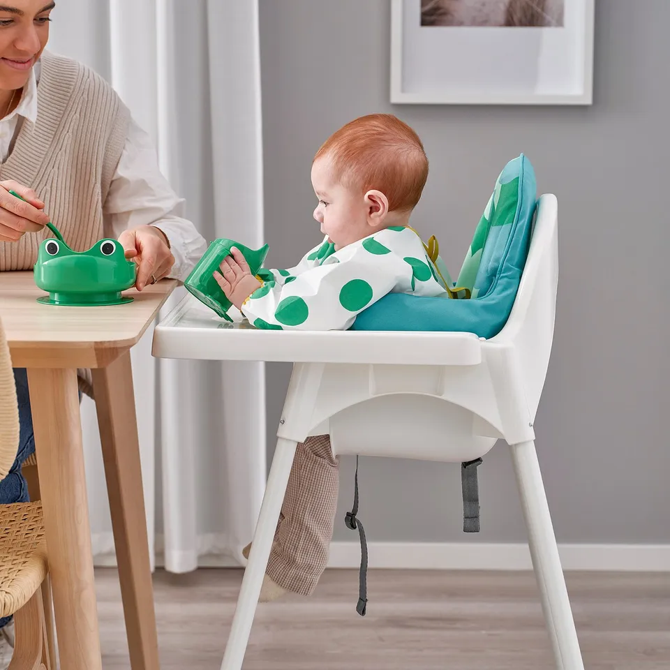 Child sits in white ANTILOP highchair, secured with a safety belt, eating from a green frog-shaped bowl held by an adult.