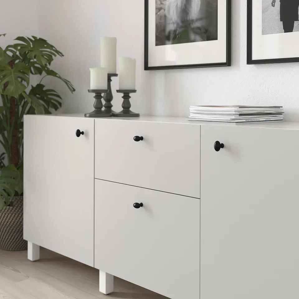 White cabinet with black knobs, holding candles and books, next to a plant.