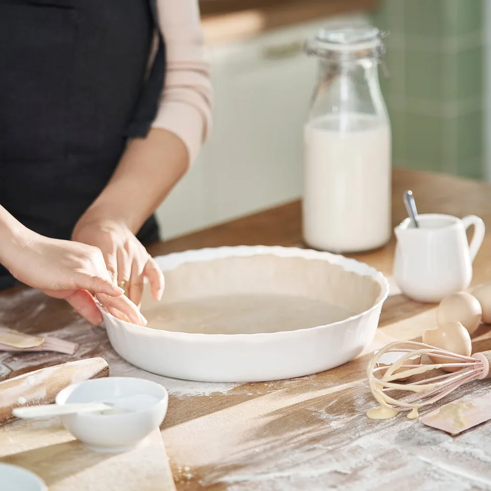 Person preparing pie dough in a white ceramic pie dish on a floured wooden table, surrounded by ingredients and kitchen tools.