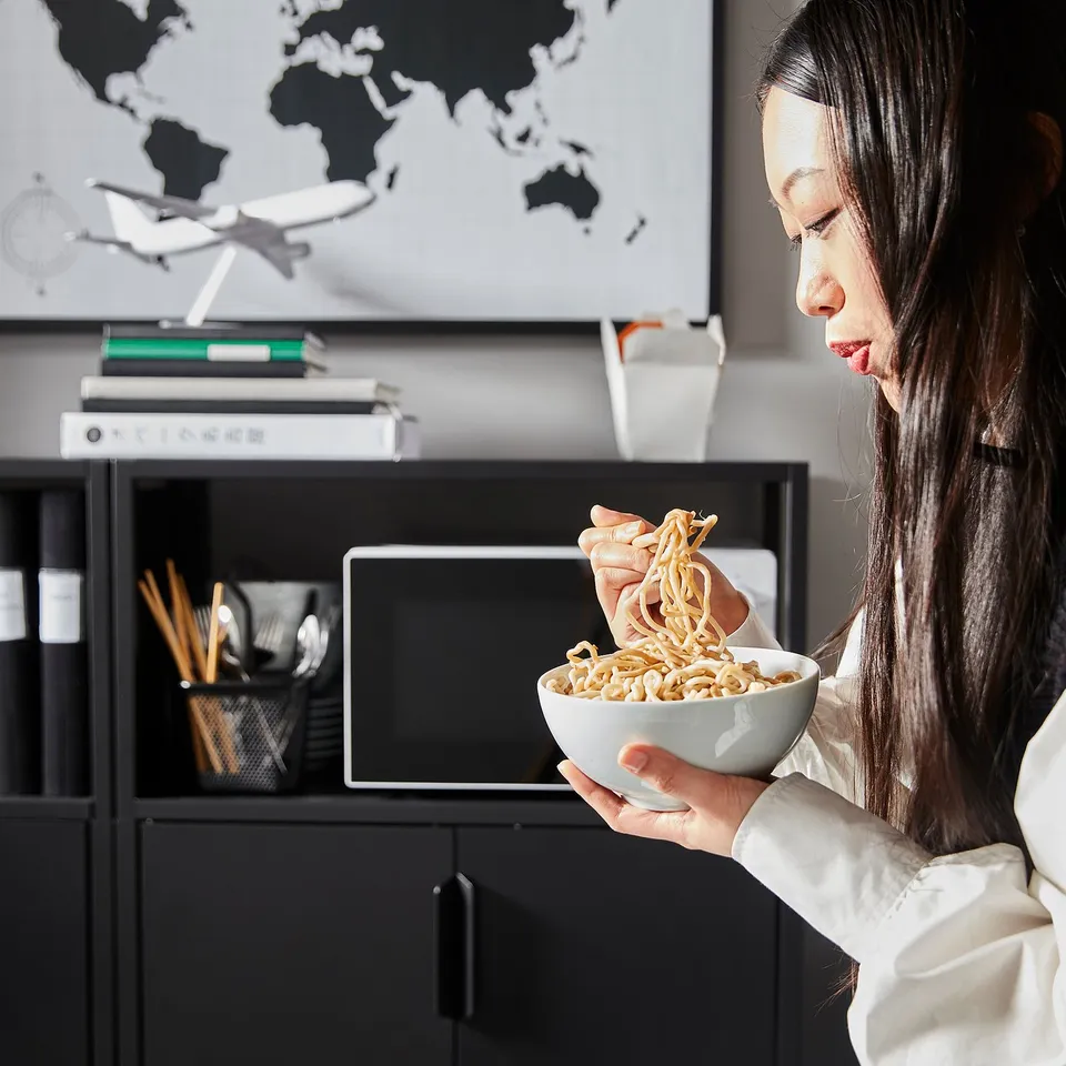 Person eats noodles at desk with computer, map, and storage.