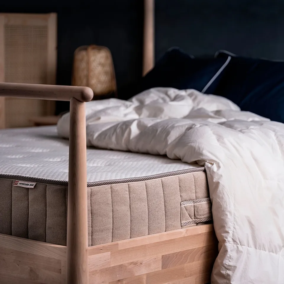 Bed with white mattress & linen cover. Dark pillows, wooden frame.