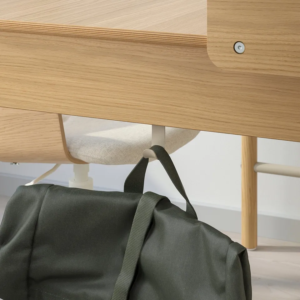 A green backpack hangs from a hook under a wooden desk, showcasing the desks cable management and privacy shield.