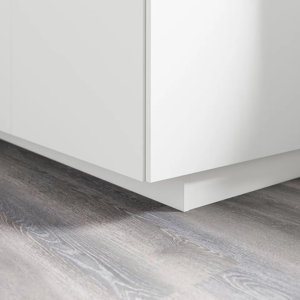 White kitchen cabinet base with a plinth covering the gap between the floor and cabinet. Grey wood-look floor visible.