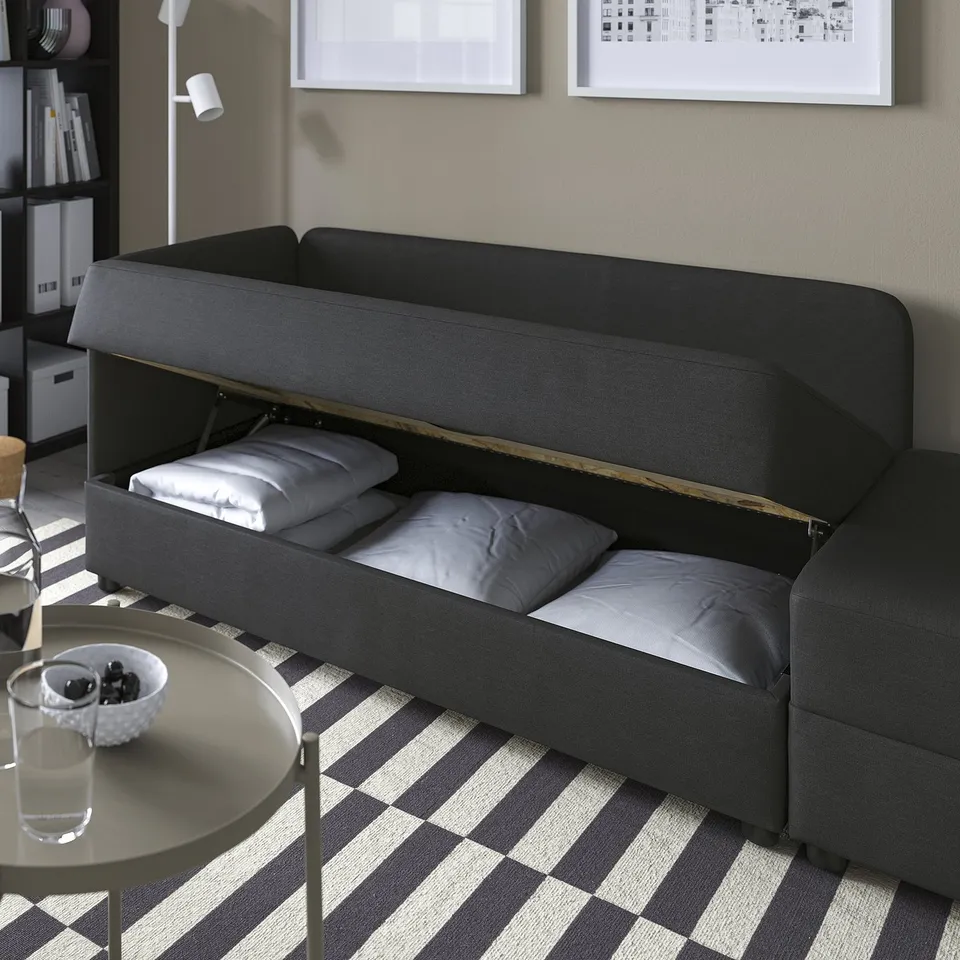 Grey sofa bed with lift-up seat revealing storage and pillows, on striped rug beside small table with glasses and bowl.
