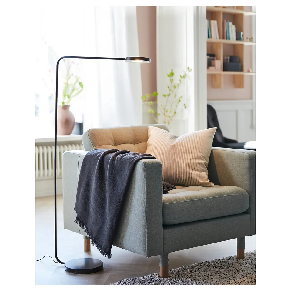 Cosy grey armchair with wooden legs, soft pillows, and a dark lamp beside it. Bright, modern living room setting with bookshelves and windows.