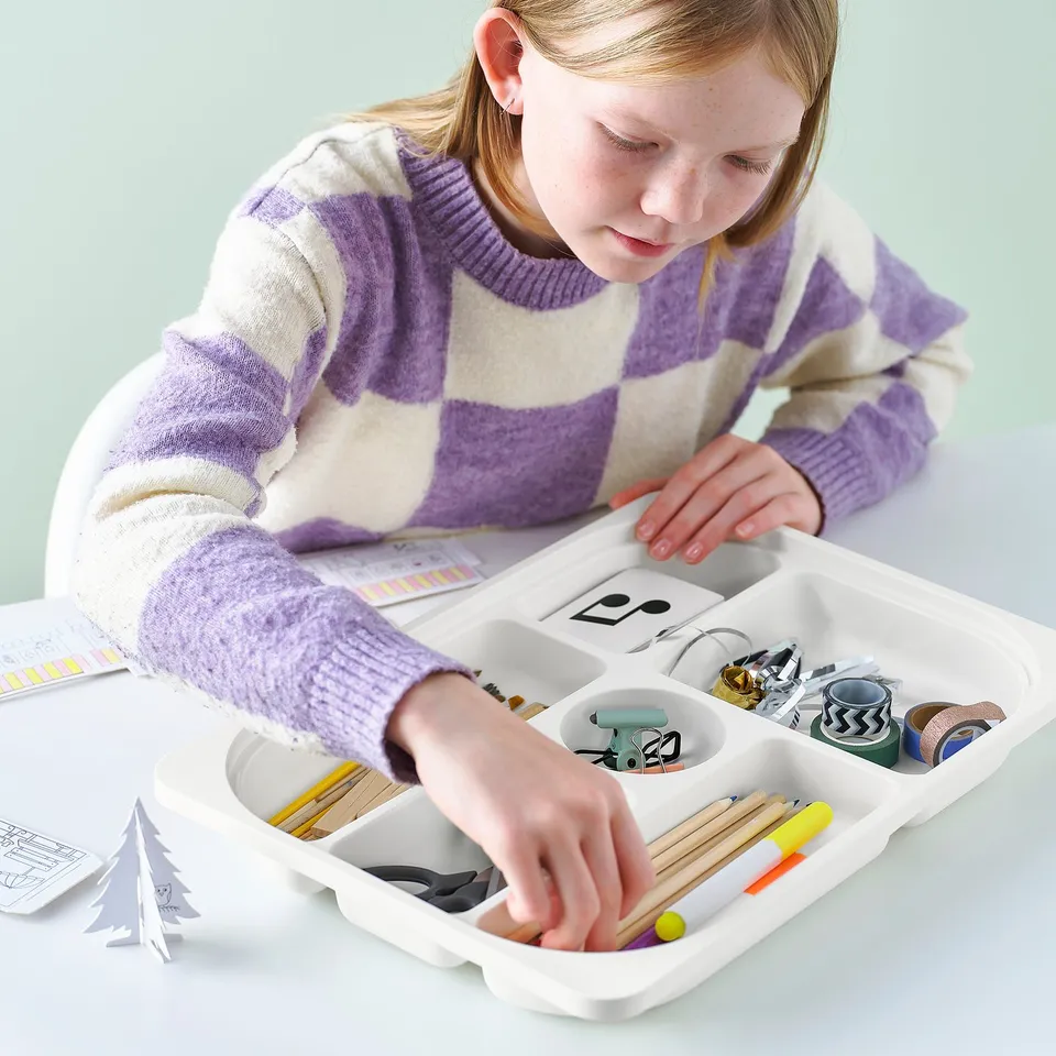 A young girl organises art supplies in a multi-compartment TROFAST tray on a table.