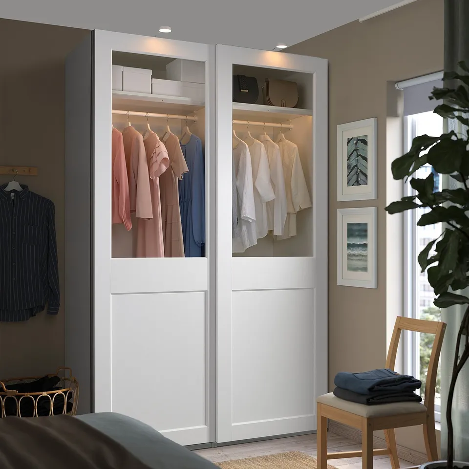 Modern white PAX wardrobe display with light, featuring glass doors and clothes racks, surrounded by furniture.