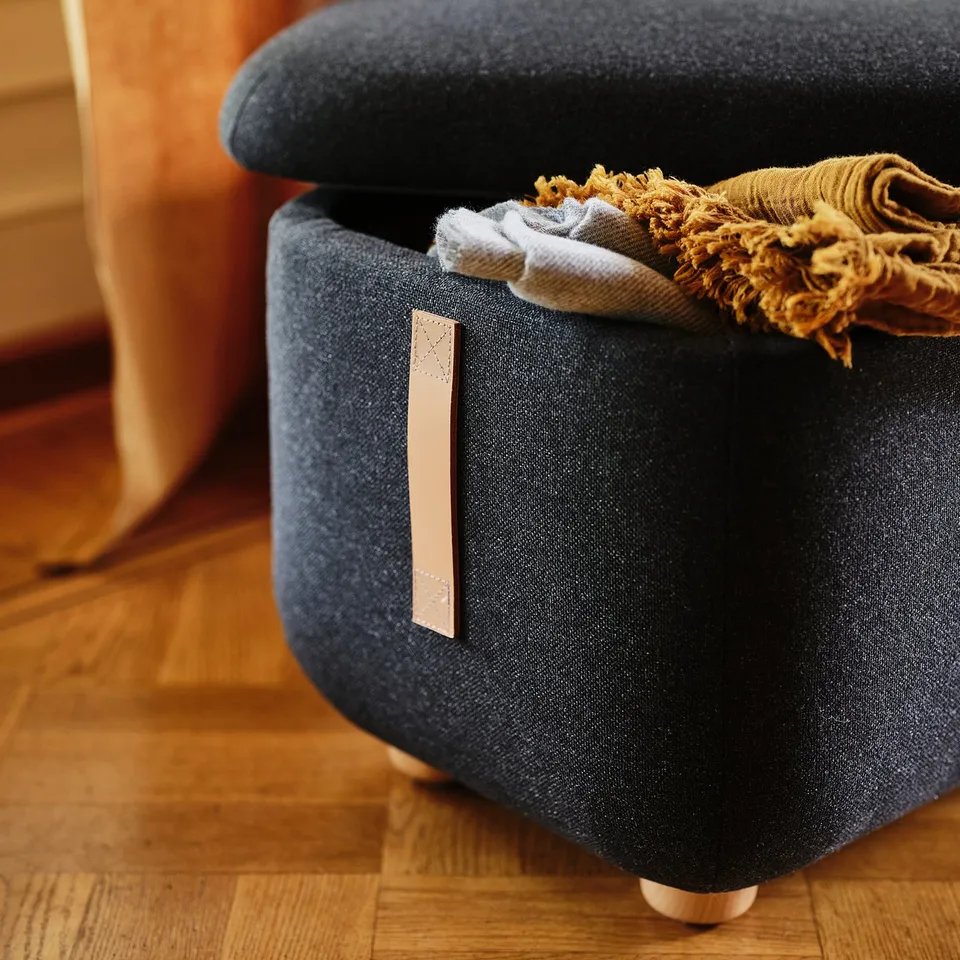 Dark grey footstool with wood legs, open lid showing folded blankets, OSKARSHAMN label.