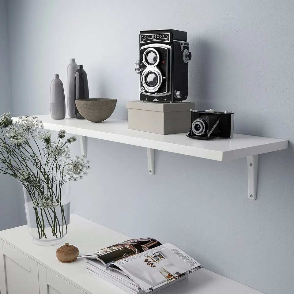 A sleek white wall shelf supports vintage cameras and modern vases, with a bevelled edge. It holds up to 20 kg, displaying items stylishly.