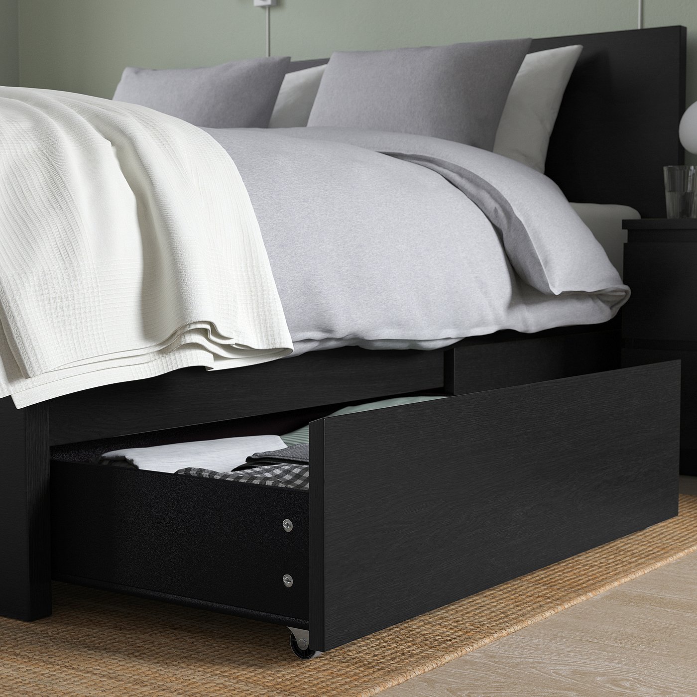 Black bed frame with drawer open, showing folded clothes. Grey bedding with pillows and white blanket.