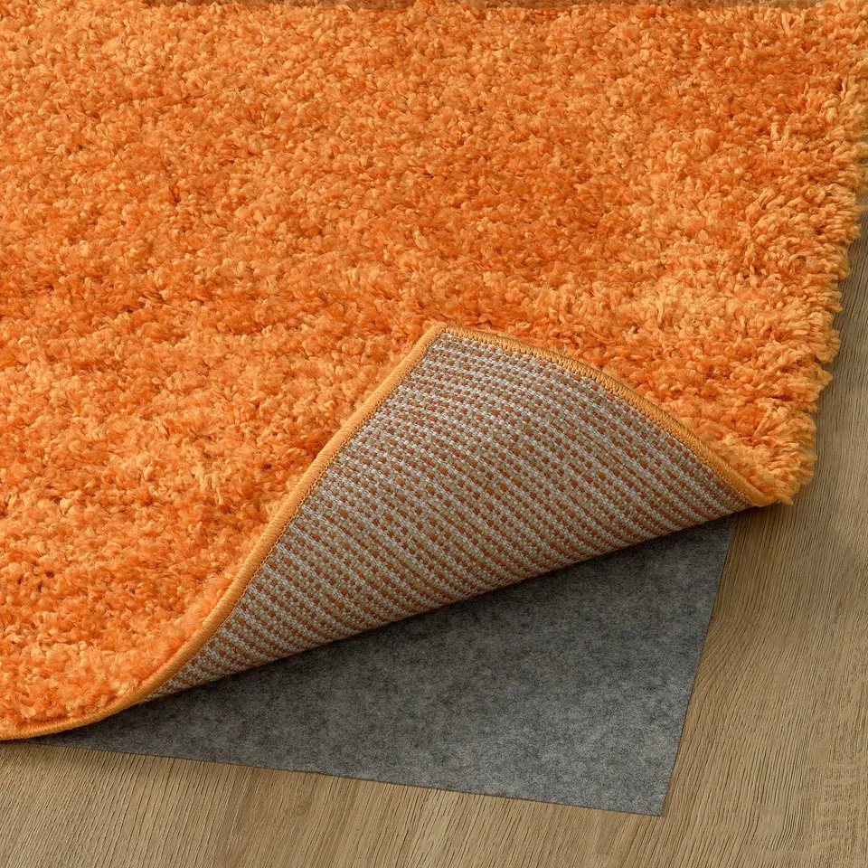 Bright orange shaggy rug with a non-slip grey mat underneath, showing its thick, soft pile designed for comfort and sound dampening.