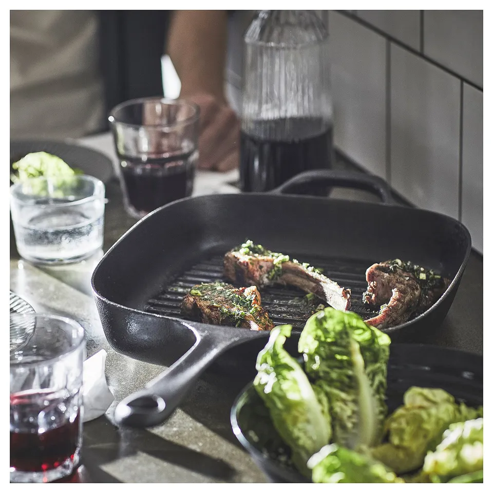 A black cast iron skillet with meat cooking on a kitchen counter, surrounded by glasses of red wine and a bowl of green vegetables.