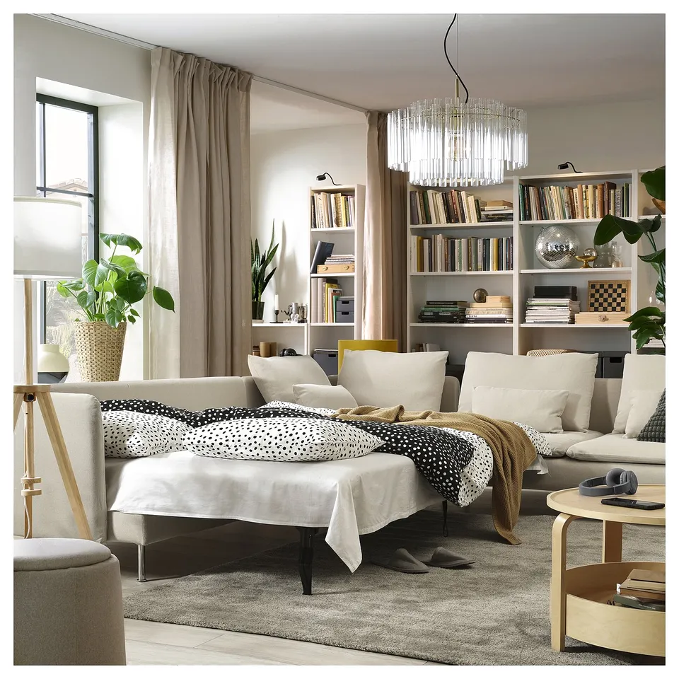 Modern living room with a beige SÖDERHAMN sofa-bed, dotted blanket, wooden coffee table, and bookshelf with white curtains and plants.