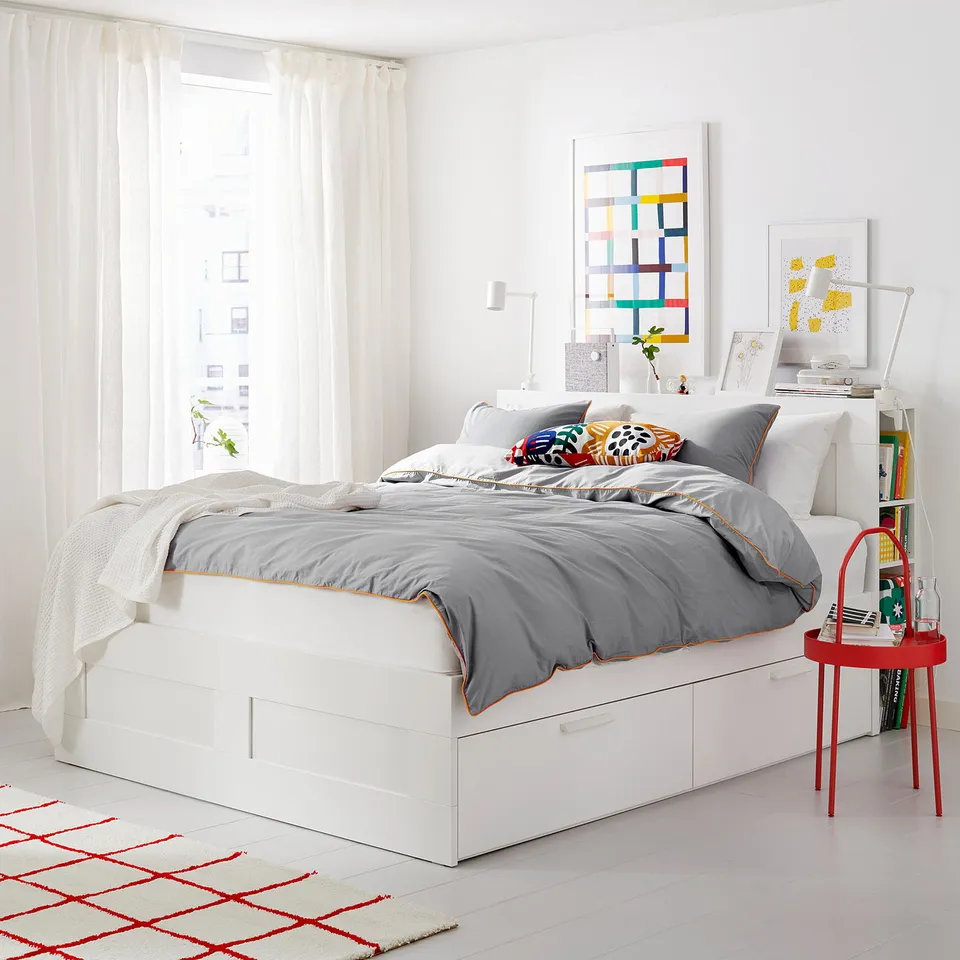 Bright, modern bedroom with white BRIMNES bed, grey bedding, colourful pillows, red side table, geometric rug, and wall art.