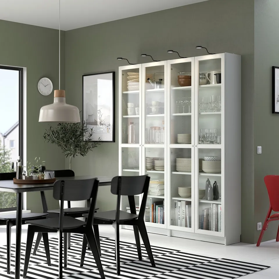 Contemporary dining room with BILLY bookcase, black chairs, striped rug, and green walls. Plants, art, and pendant light adorned.