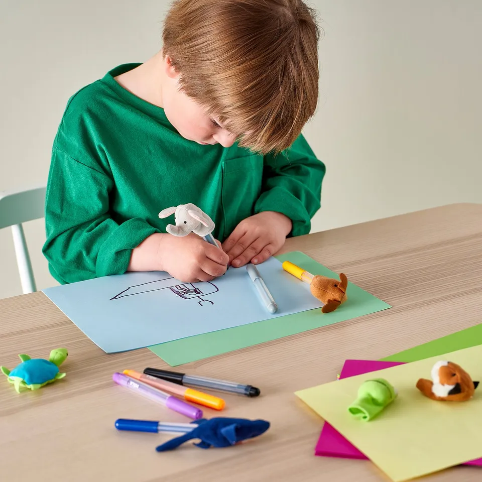 Child draws with MÅLA art supplies at wooden table.