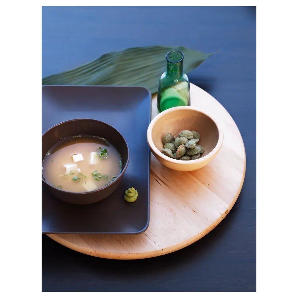 A wooden tray holds a blue bowl of soup and a bowl of green sweets, with a green bottle beside.