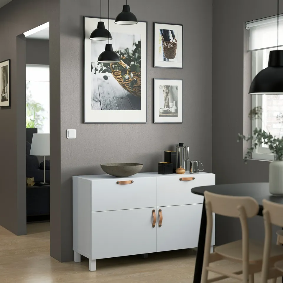 Minimalist white BESTÅ sideboard with light wood handles, black wall lamps, and framed art in a modern dining space.