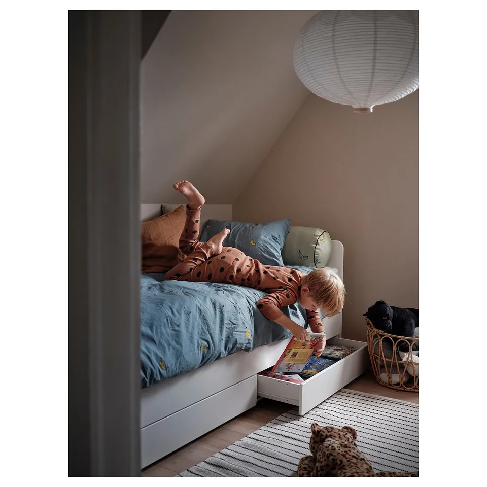 Child playing under a white SLÄKT trundle bed with light blue bedding and a hanging lamp.