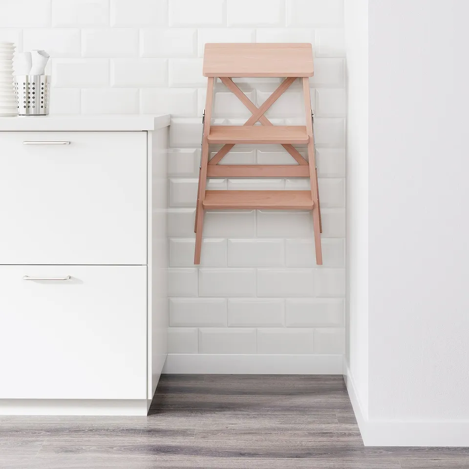 A foldable wooden step ladder leans against a white tiled wall in a kitchen. Its designed for reaching high cabinets and folds to save space.