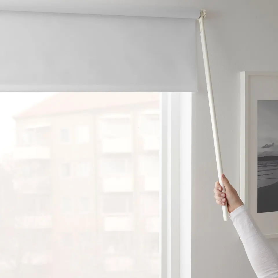 A hand pulls a long, white roller blind wand, adjusting a light-blocking fabric over a large window. The wands design enables precise positioning.