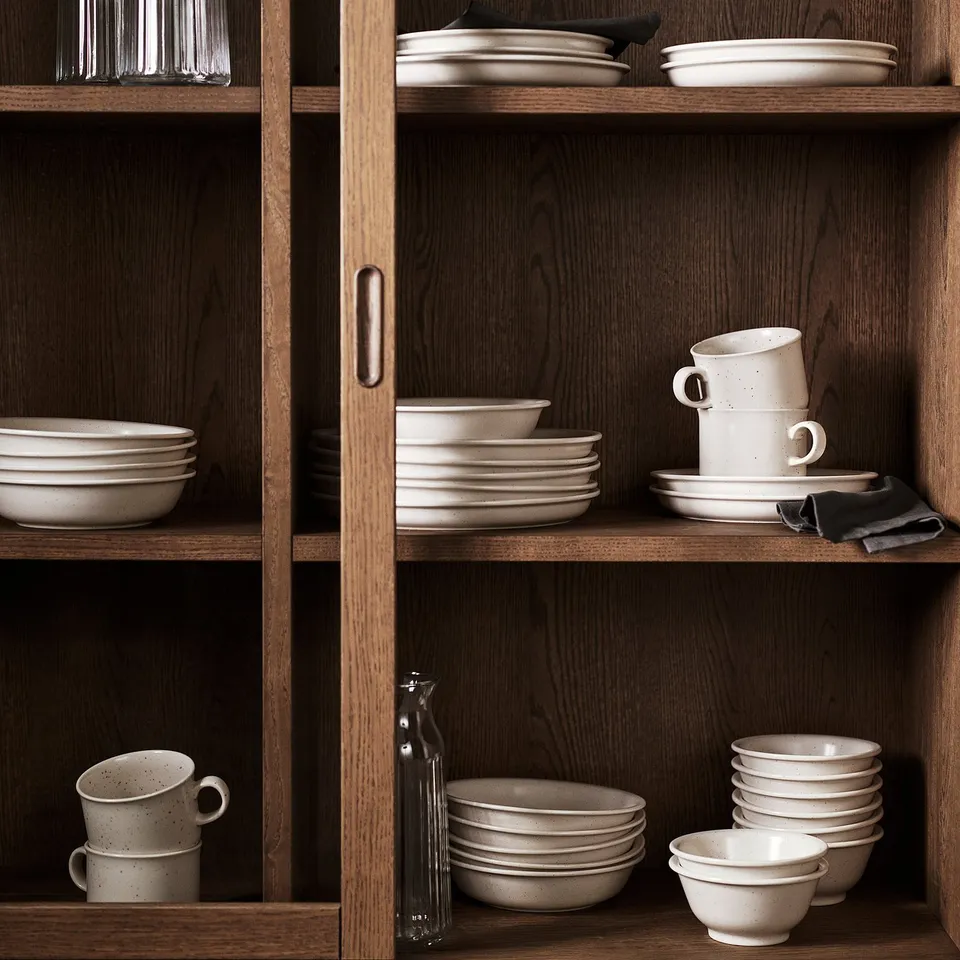 Wooden cupboard with DAGGASTER stoneware in light beige, including stacked mugs, plates, and bowls of varying sizes.