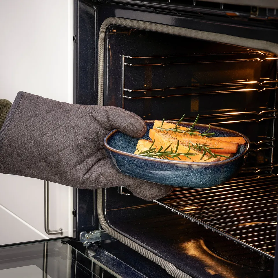 Person wearing oven mitts places blue ceramic dish with cooked food into oven, highlighting GLADELIG oven-to-table functionality.