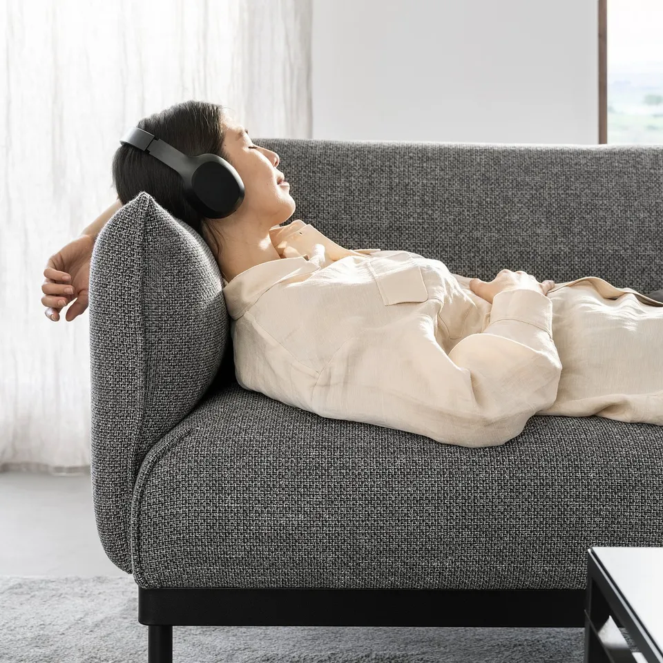 A person relaxes on a grey ÄPPLARYD sofa, wearing headphones, with beige cushions, in a calm living room setting.