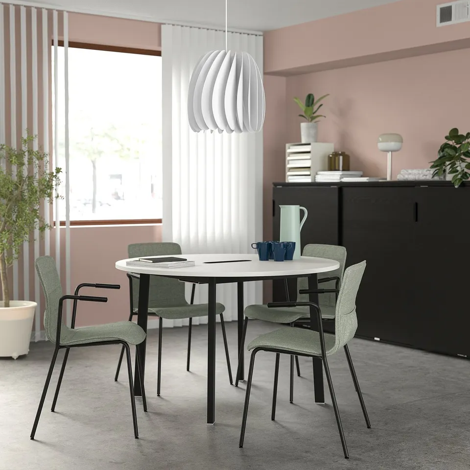 Modern round white MITTZON conference table with green chairs by window. Black cabinet with books and a blue mug. Small, ideal for small groups.