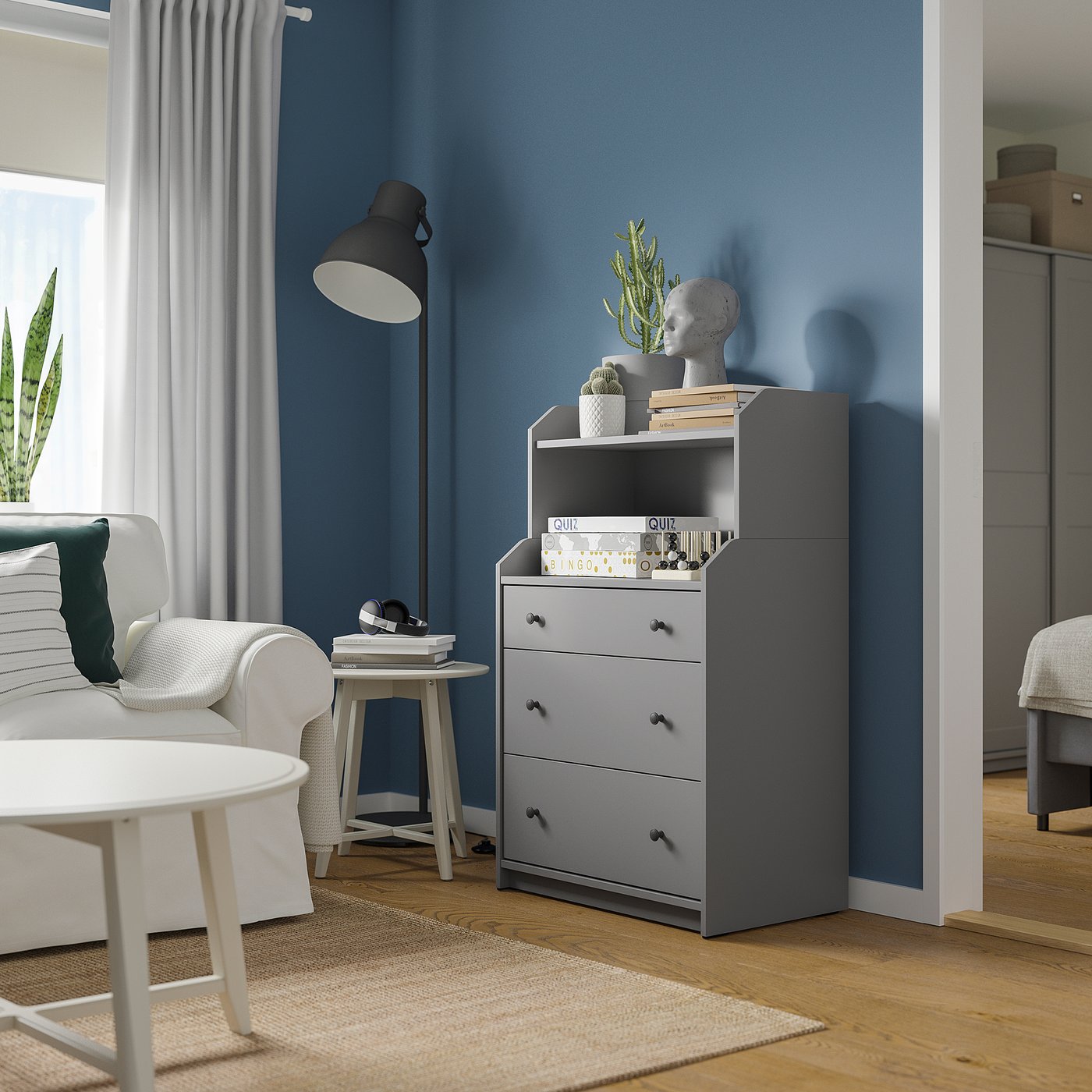 A modern living room with blue walls, a white couch, and a grey HAUGA chest. It has shelves with books and a sculpture.