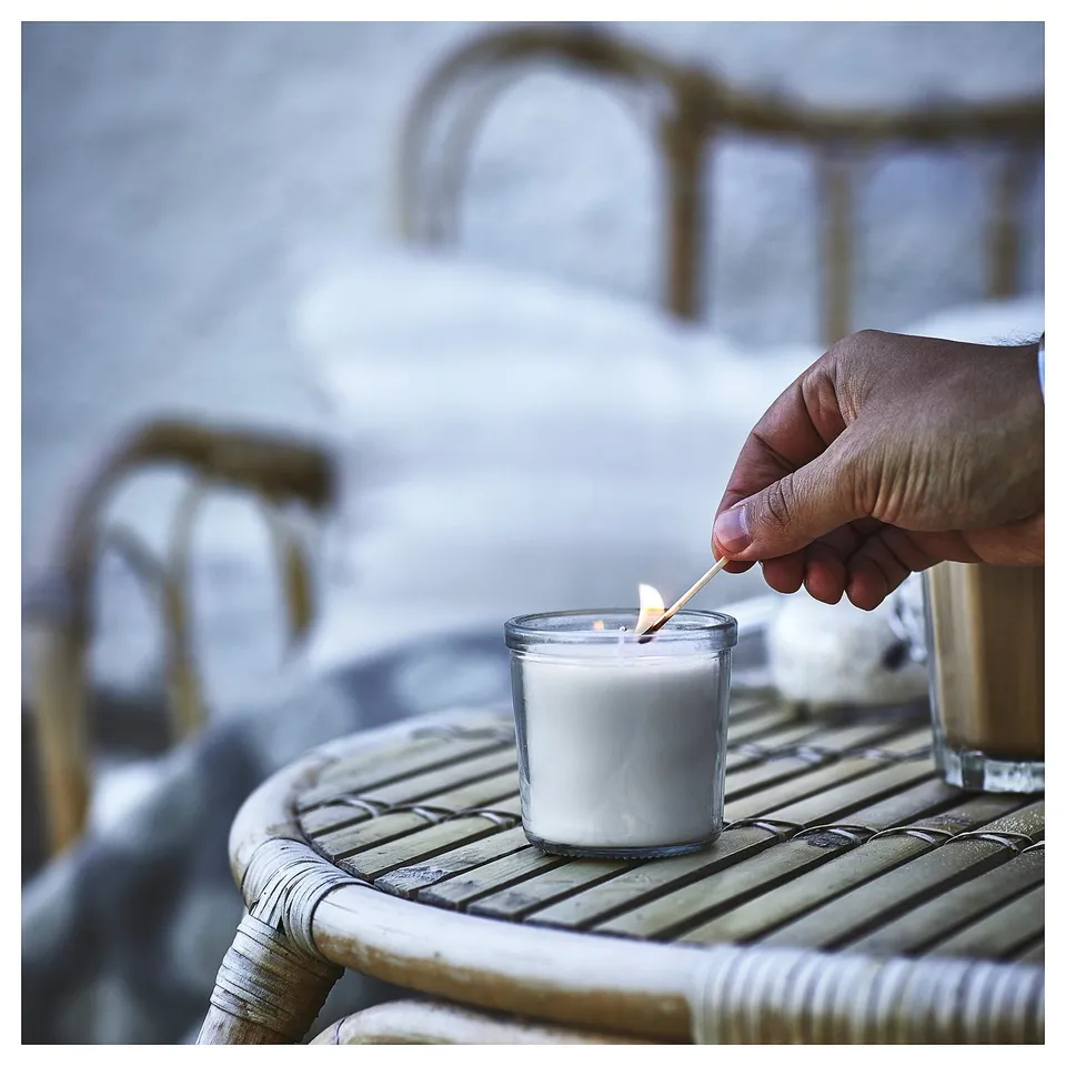 A hand lights a white JÄMLIK candle in a glass holder on a wicker table, creating a cosy atmosphere.