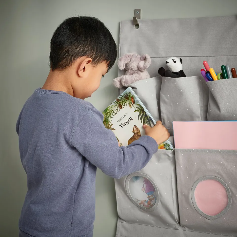 Child reaching into grey hanging organiser with pockets, placed on a wall. The organiser holds books, toys, and art supplies.
