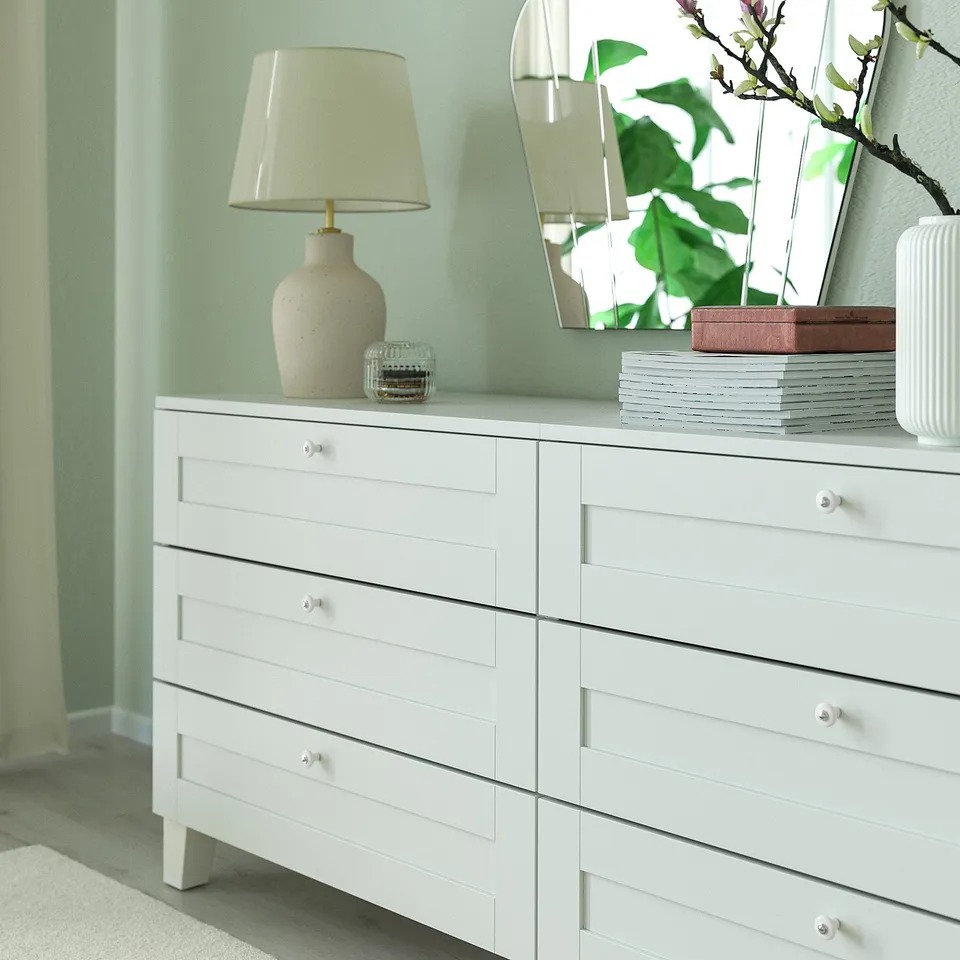 White dresser with six drawers against light green wall. Decorated with lamp, mirror, and vase.