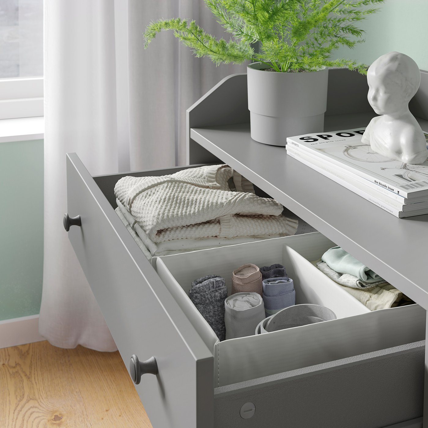 Open grey HAUGA dresser with folded clothes and socks, next to a white bust statue and green plant.