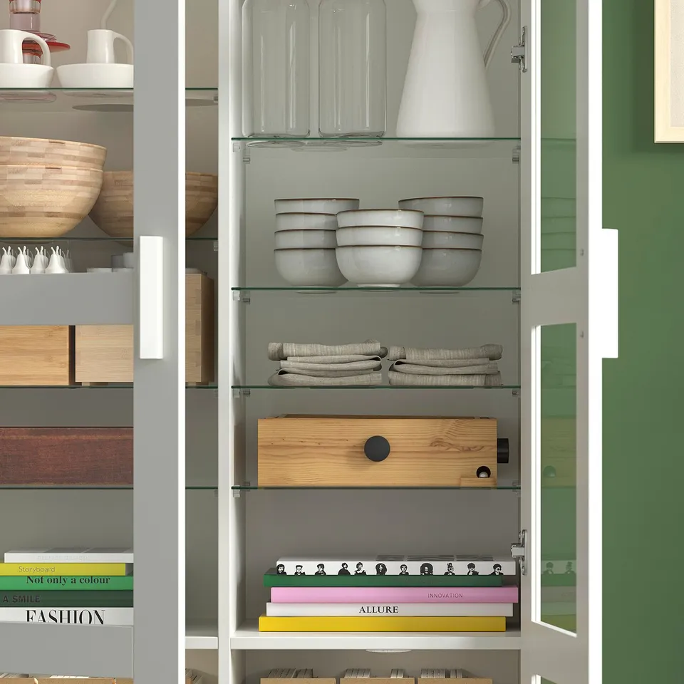 A modern kitchen cabinet with glass shelves, storing bowls, jars, pitchers, towels, a wooden box, and books.