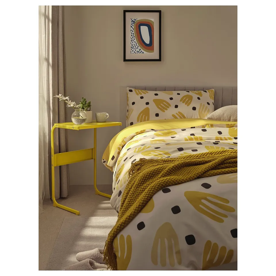 Bedroom scene with yellow bedding, yellow side table, patterned duvet, pillow, art on wall, curtains, vase, and cup.