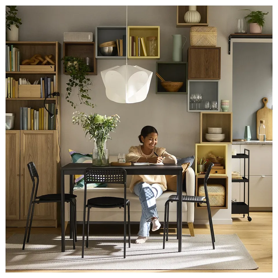 Young girl sitting at table with laptop, surrounded by decorative shelves with items. Shes wearing a brown jacket, blue jeans, and trainers.