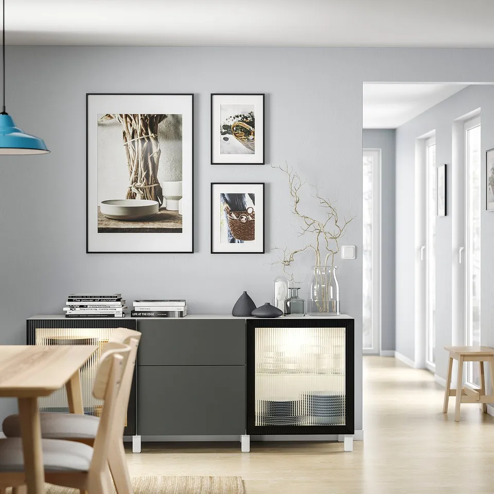 Modern dining area with light wood table, chairs, and grey BESTÅ storage unit featuring clear glass doors, black accents, and art prints on wall.