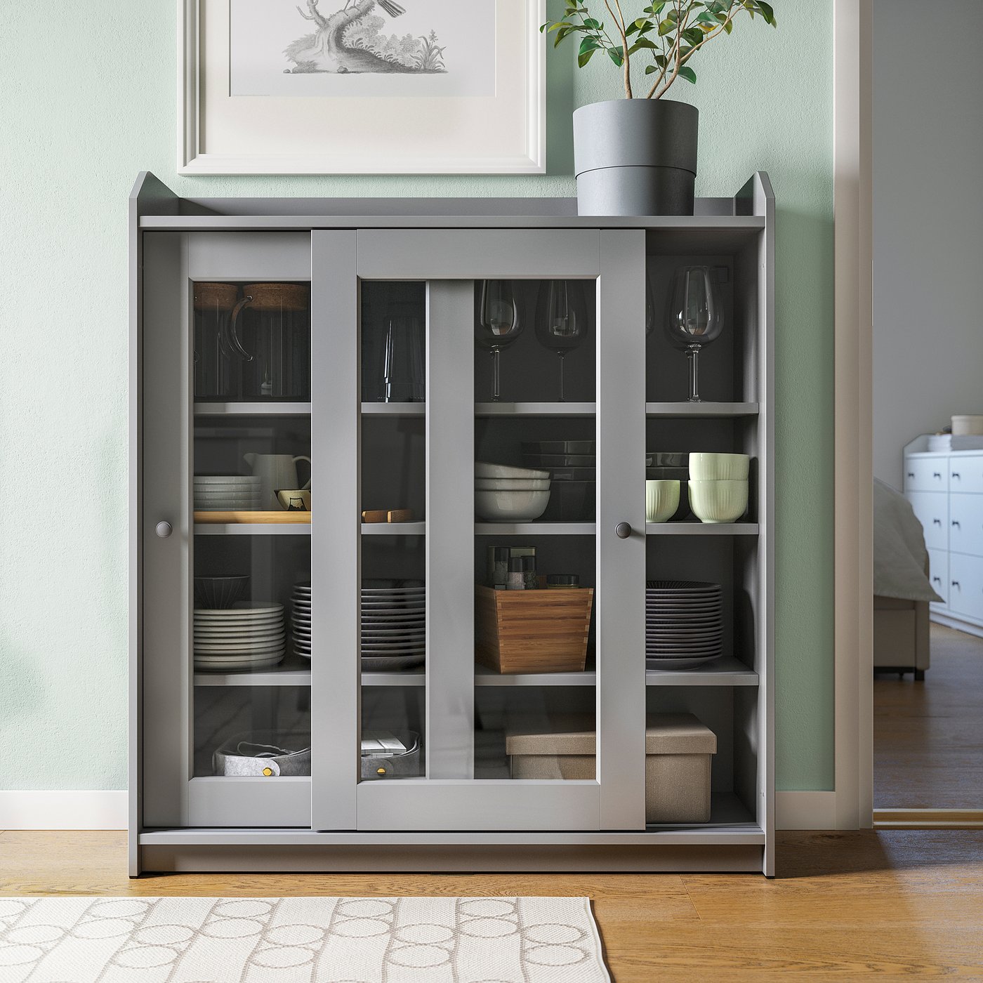 Grey HAUGA display cabinet with glass doors and open shelves, storing dishes, bowls, and glasses in a living room setting.