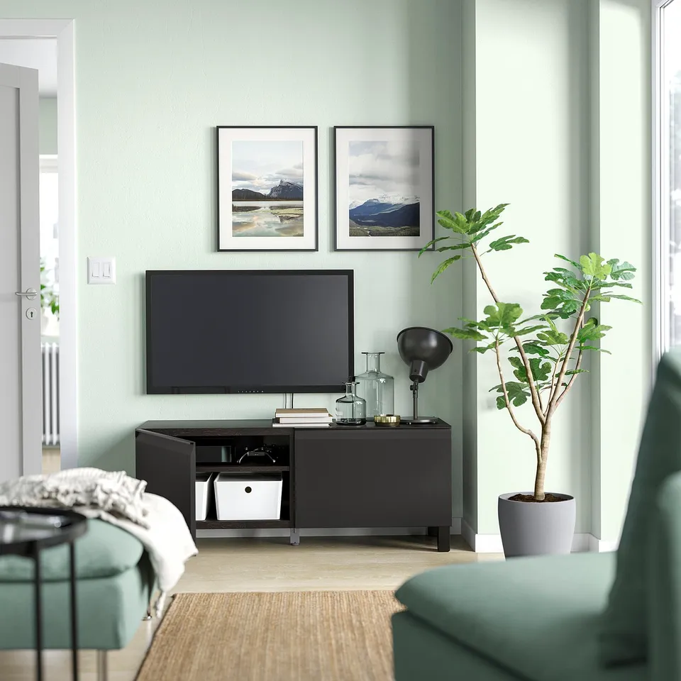 Modern living room with green walls, black BESTÅ tv stand, framed mountains, plant, teal chair.