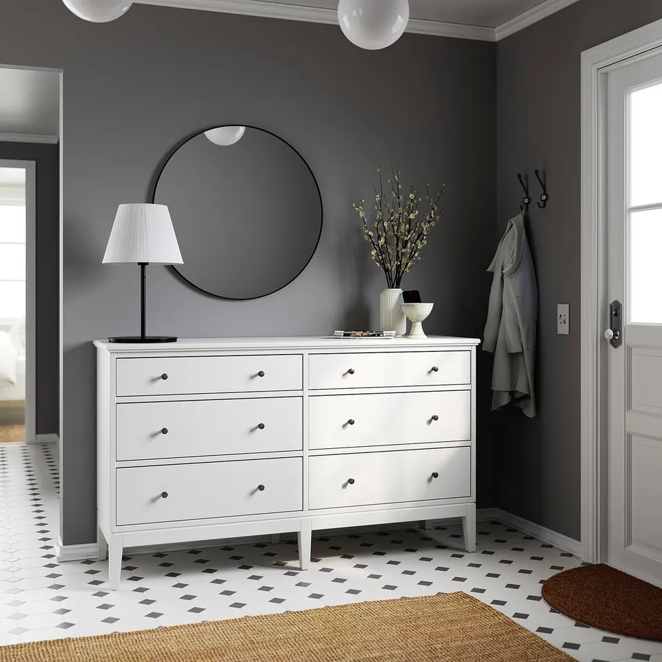 Modern entryway a white IDANÄS chest with 6 drawers, rounded mirror, lamp, vase with dried flowers, coat hooks, and patterned tile floor.