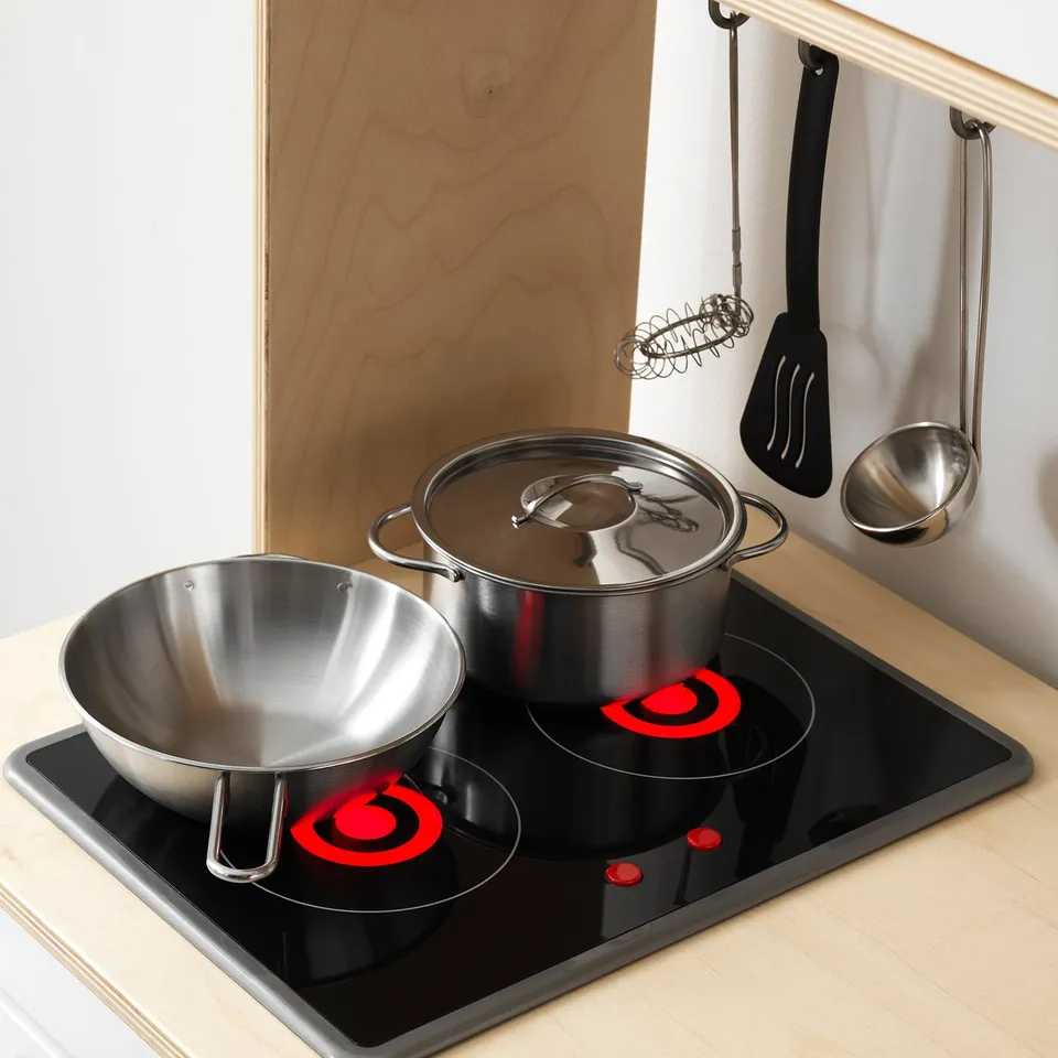 Stainless steel play pots and pans on a black toy stove with red heat indicators, mocking real cooking.