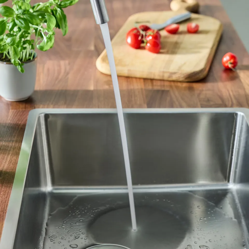 Kitchen sink with modern mixer tap running water, wooden cutting board, tomatoes, and basil plant.