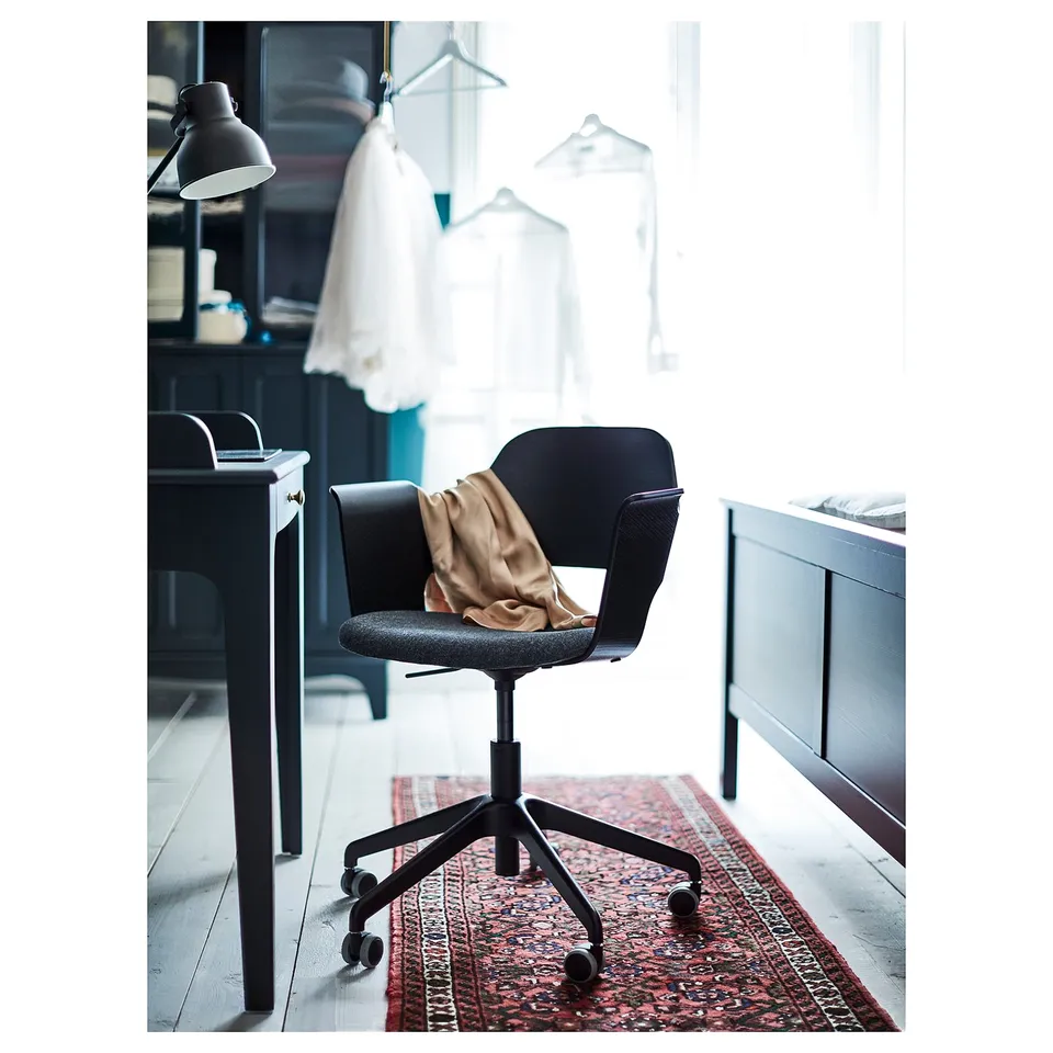 Modern black office chair with wheels, gold throw, wooden desk, red patterned rug, lamp and window.