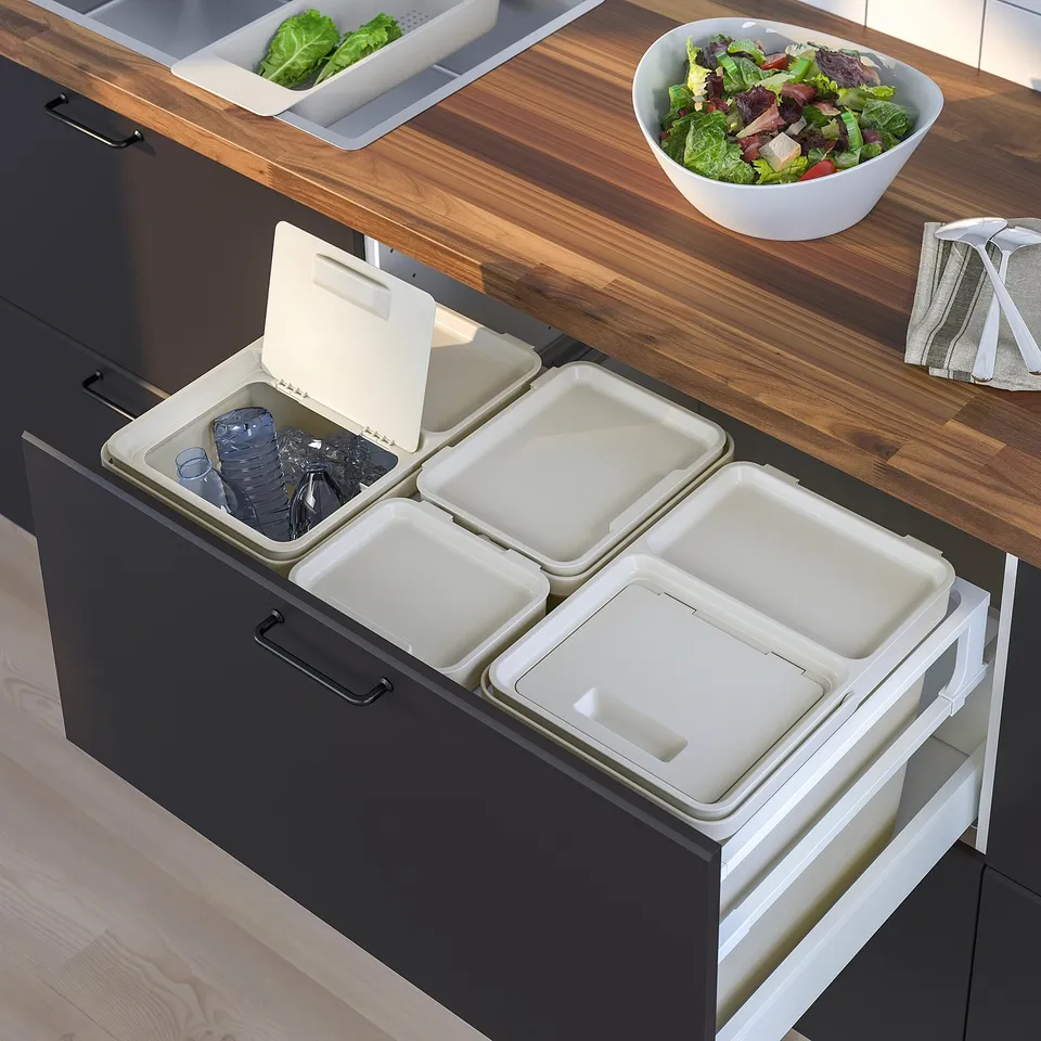 Four grey waste sorting bins under a kitchen counter, each with a lid and one open showing plastic containers. Wooden countertop with a salad bowl.