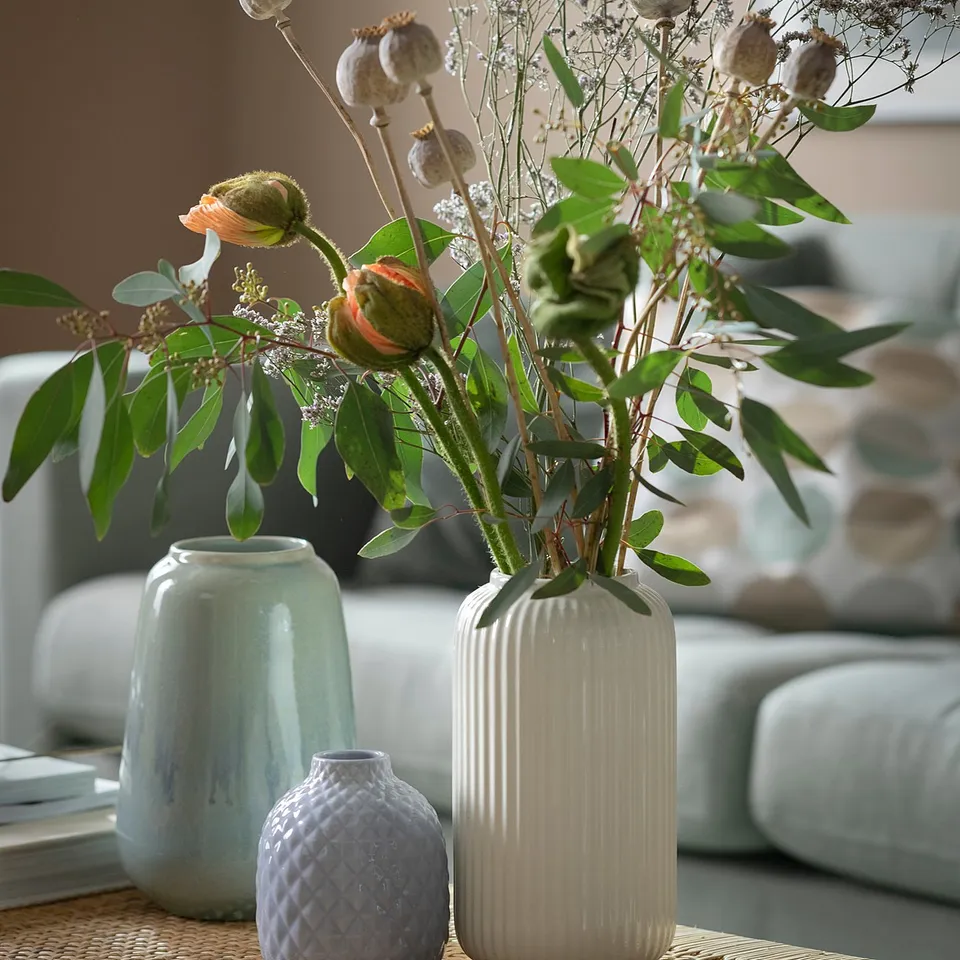 Three vases on table: large white ribbed, light blue textured, green, bud-filled.
