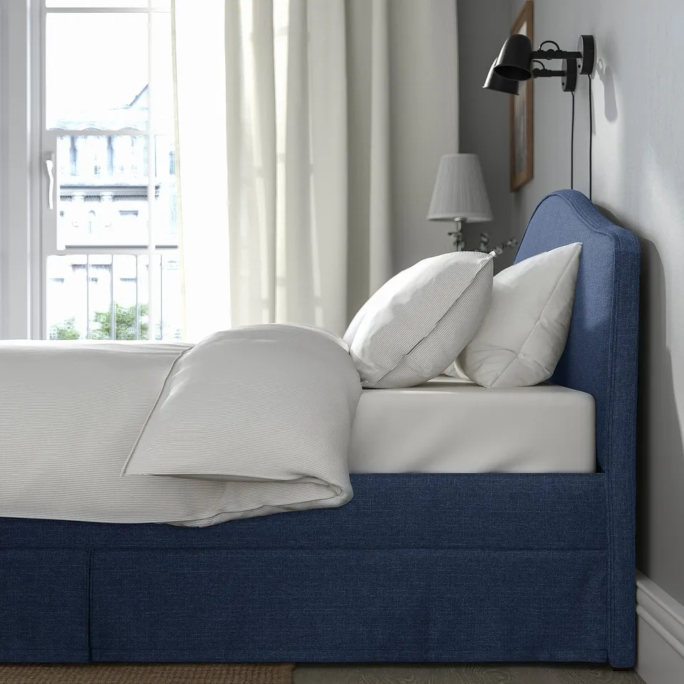 A cosy bed with a blue headboard, white bedding, and a wall lamp. The headboard is curved and padded, perfect for reading against.