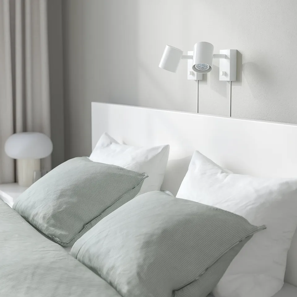 Minimalist bedroom with white bed, green sheets, dual wall lamp, mushroom-shaped bedside table lamp.