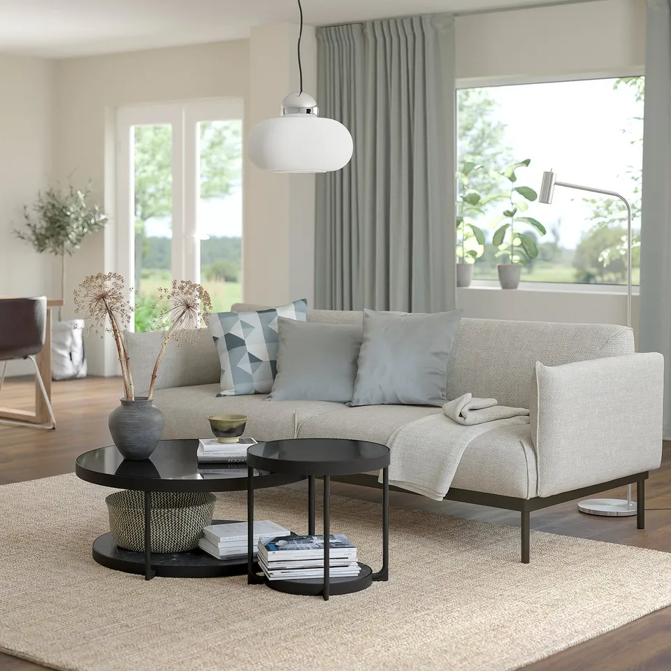 Modern living room with white sofa, grey pillows, black tables, plants, and large window.